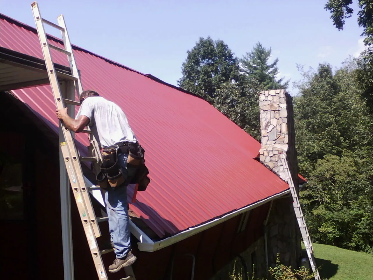 Roofer on a ladder beside a red metal roof and stone chimney — Soffit & Fascia Repair by Roofing Brothers in Sarver